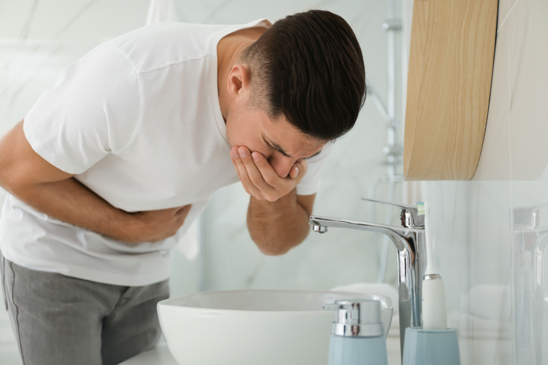 Man bending over a sink covering his mouth, indicating nausea or discomfort from a stomach flu or virus.