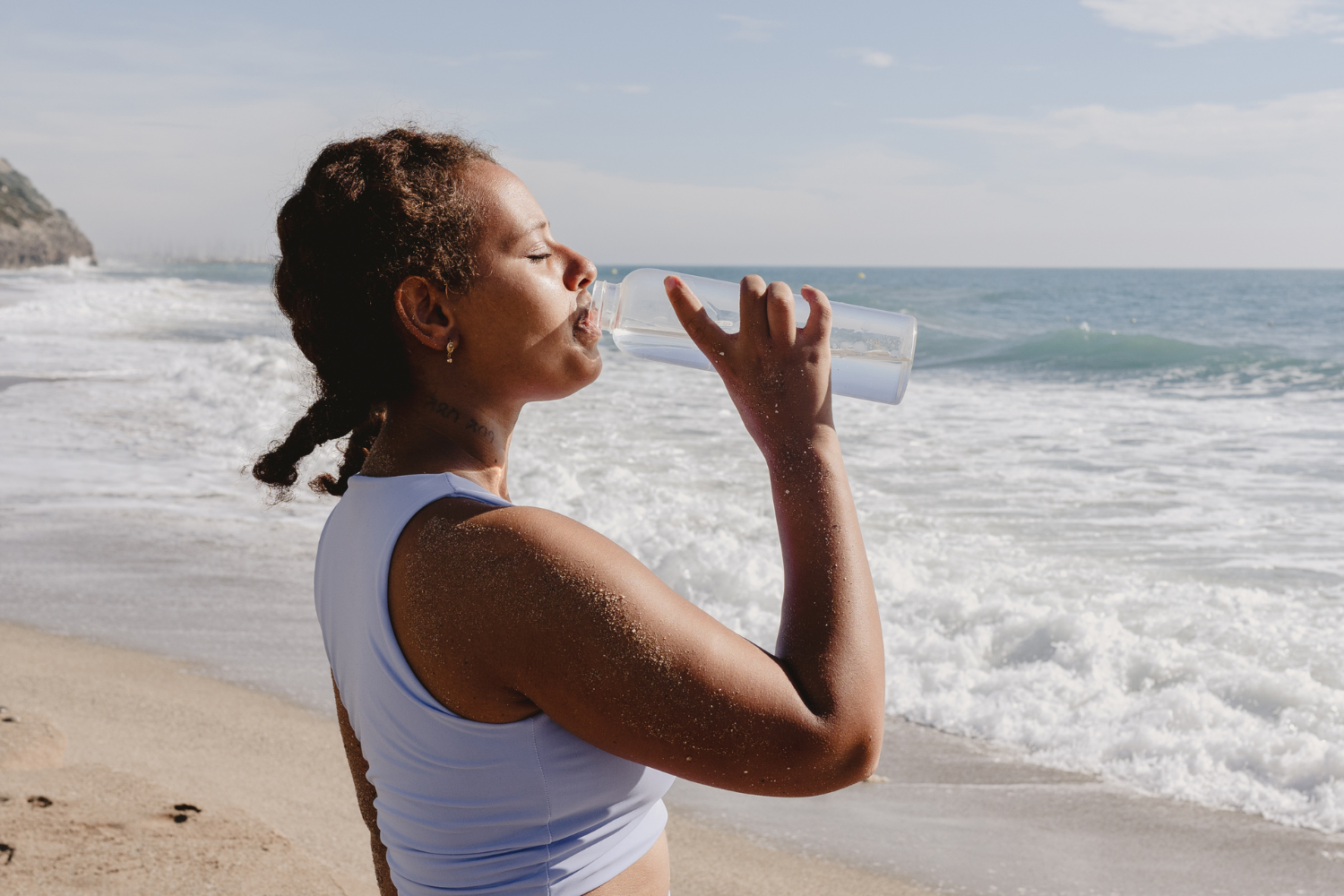 A person drinks water from a clear bottle on a sandy beach with ocean waves.