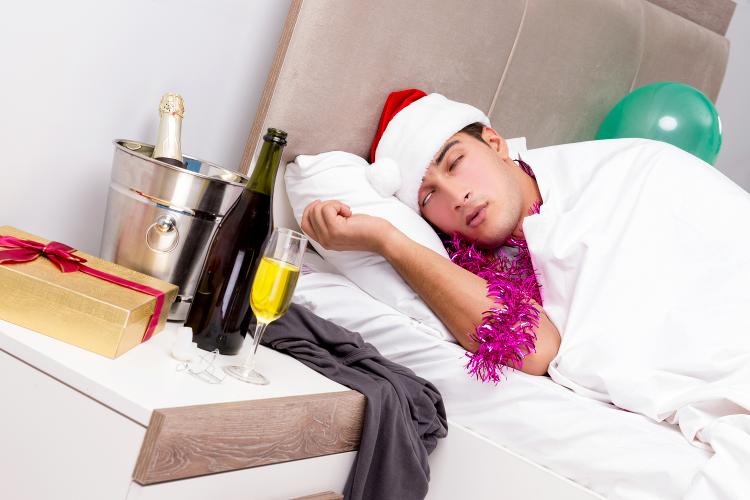 Man wearing a Santa hat sleeping in bed next to a champagne bottle, a glass of yellow liquid, and a present, depicting sleeping off a holiday hangover.