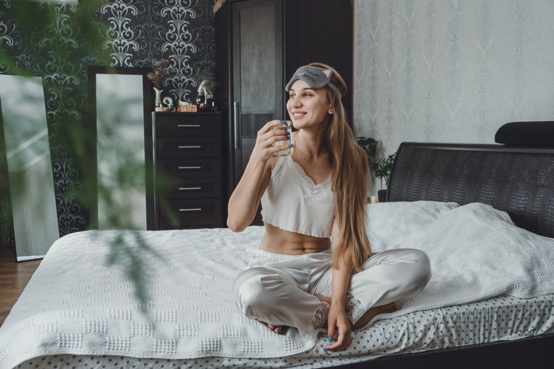 A young woman in white silk pajamas sitting cross-legged on a bed, smiling and taking the first sip of water upon waking up to begin her morning hydration routine.
