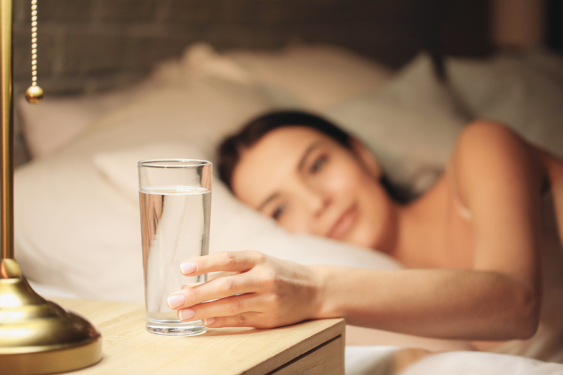 A woman waking up in bed and reaching for a glass of water on the nightstand, illustrating nighttime thirst.